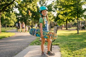 Child wearing a protective skate helmet holding a skateboard in a sunny park.